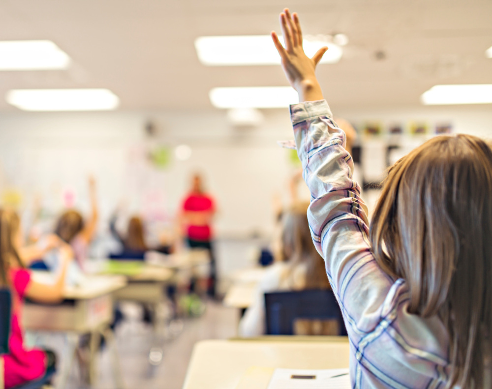 An elementary school girl raises her hand in the back of the classroom. The camera is focused on the girl and the rest of the classroom is shown blurry.
